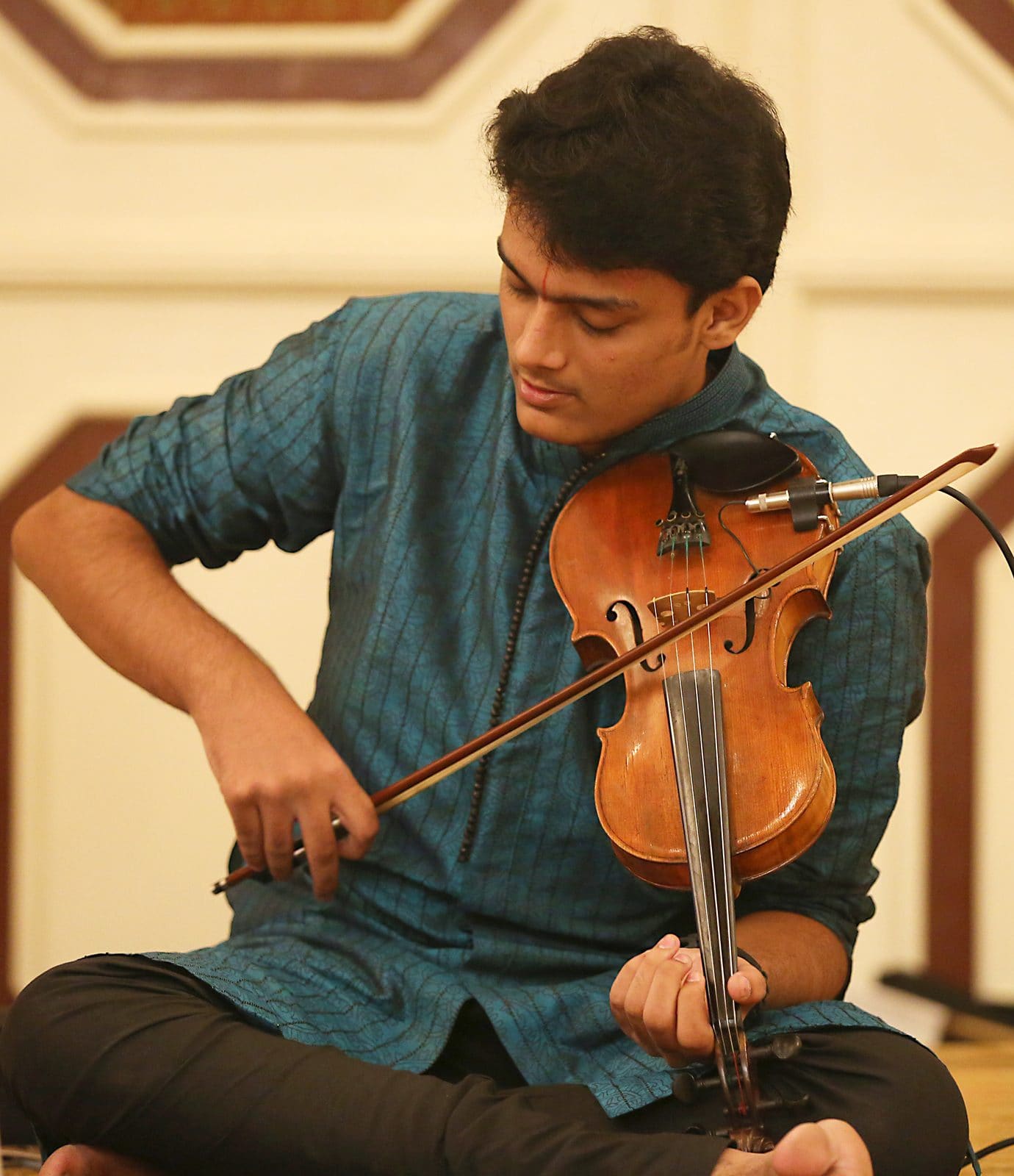 Raghavasimhan seated cross-legged on stage, playing the violin in the Carnatic posture at a concert