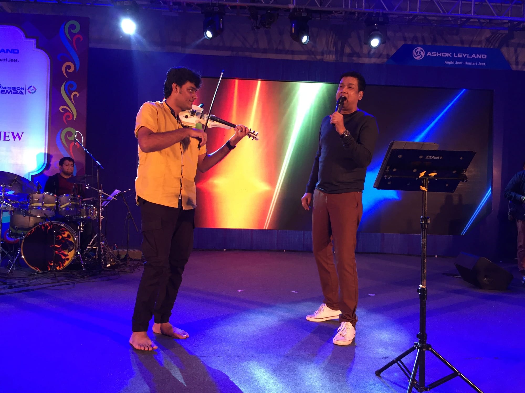 Playing violin beside a singer on a concert stage lit in blue, a rainbow screen behind