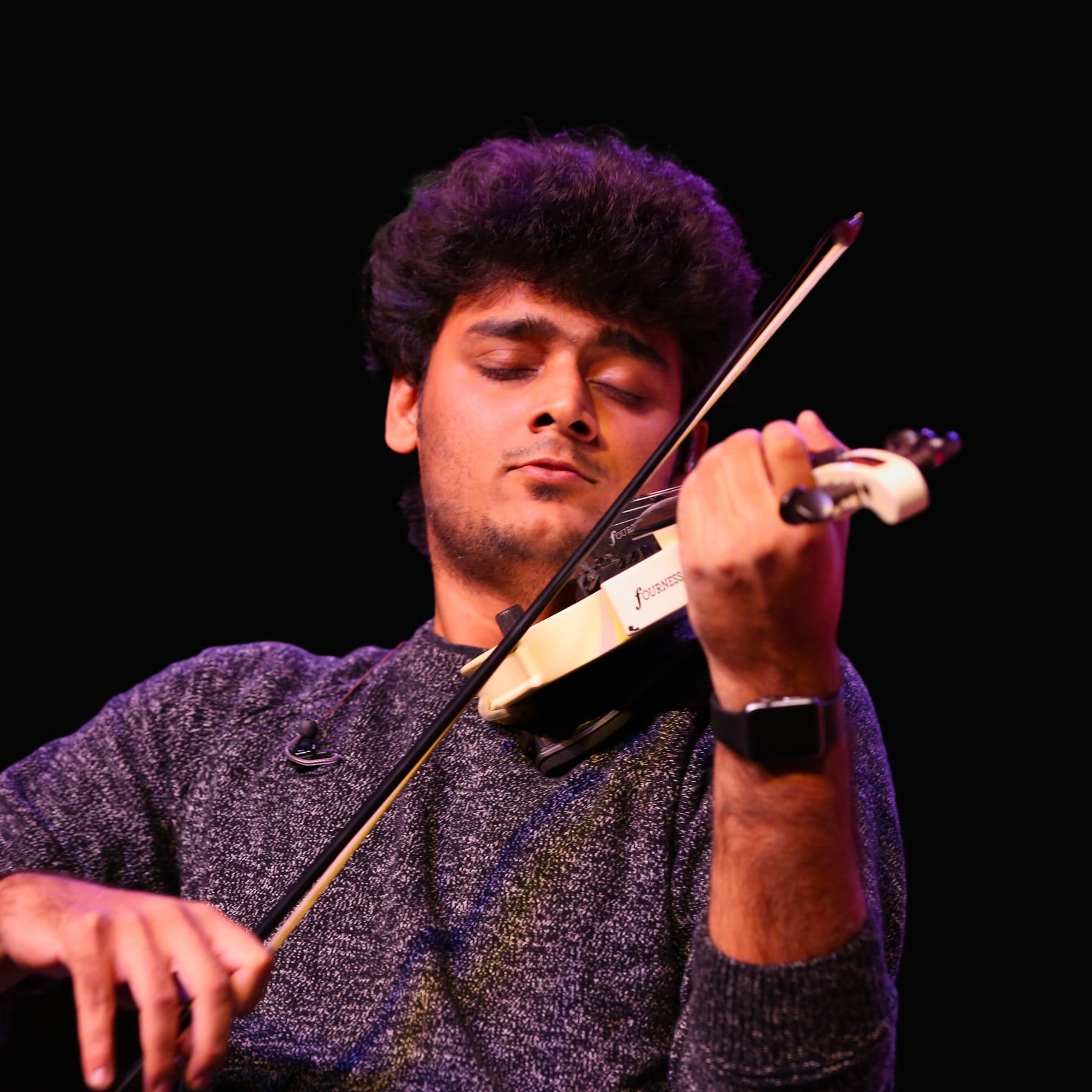Raghavasimhan, eyes closed, playing a white electric violin against a dark stage