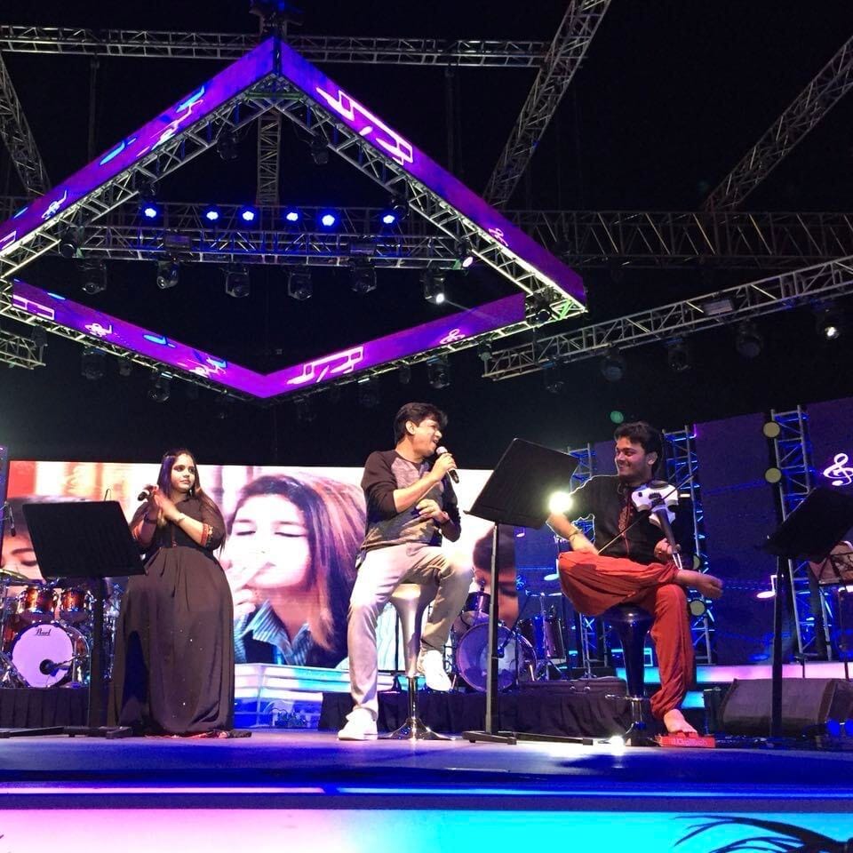 Seated with the violin on a concert stage under a lighting rig, beside two singers