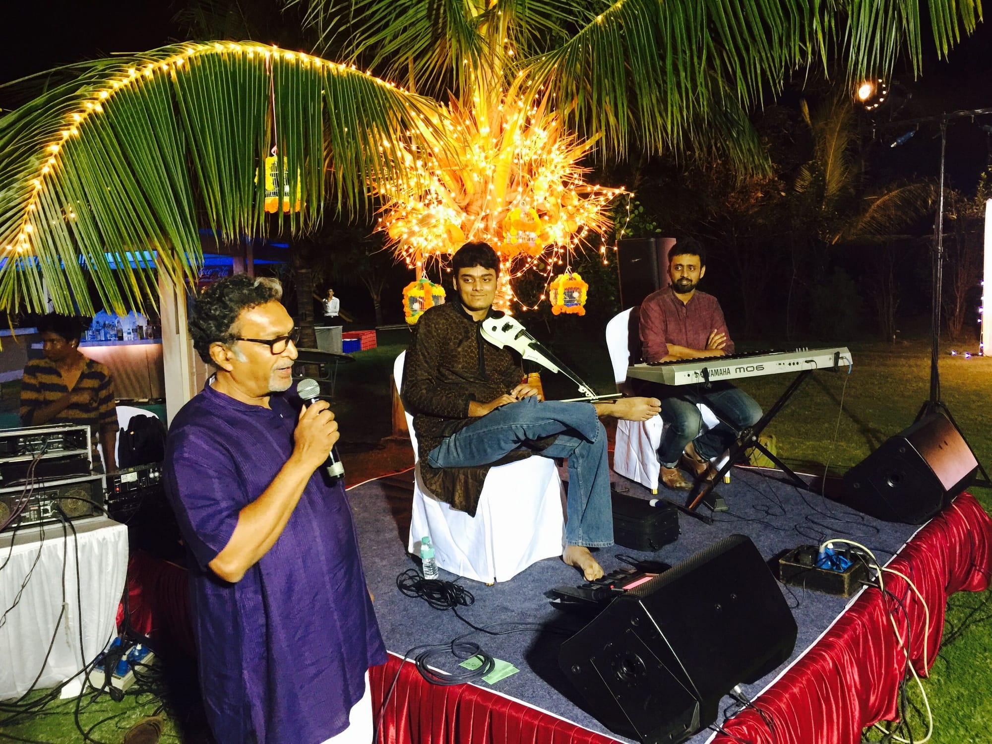 Playing a white electric violin at an evening garden concert under palm-leaf lights, a speaker at the microphone