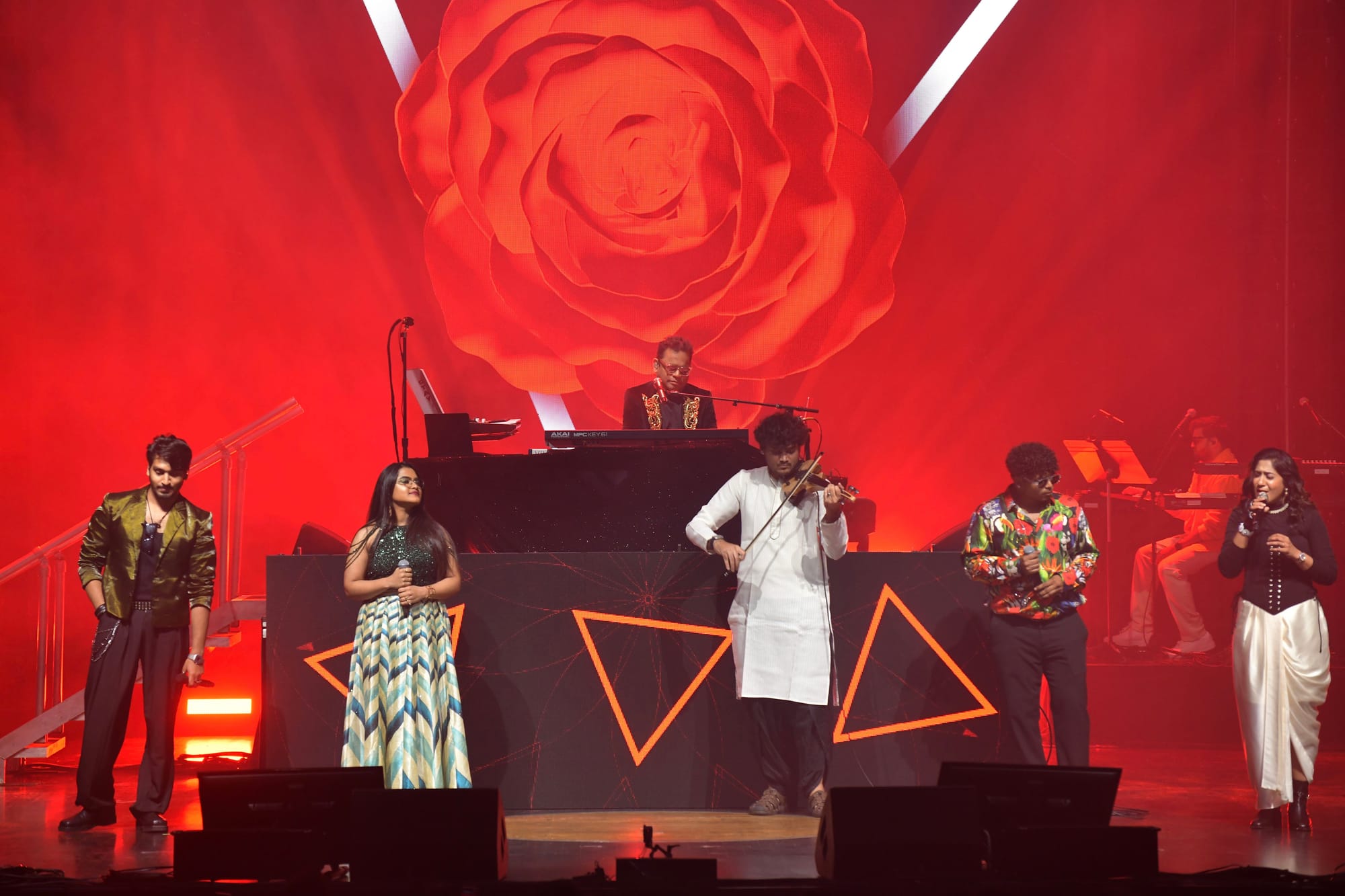 Playing violin at centre stage before a giant red rose backdrop, singers and band around him