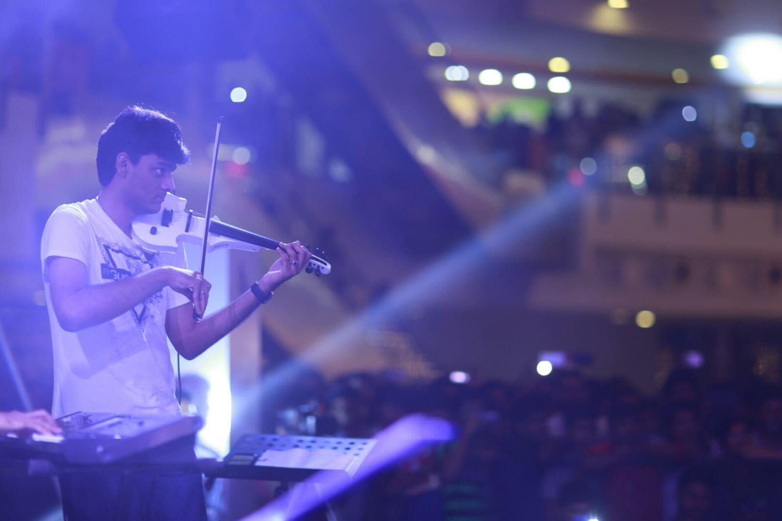 Playing a white electric violin on a concert stage under blue lights, keyboards beside him